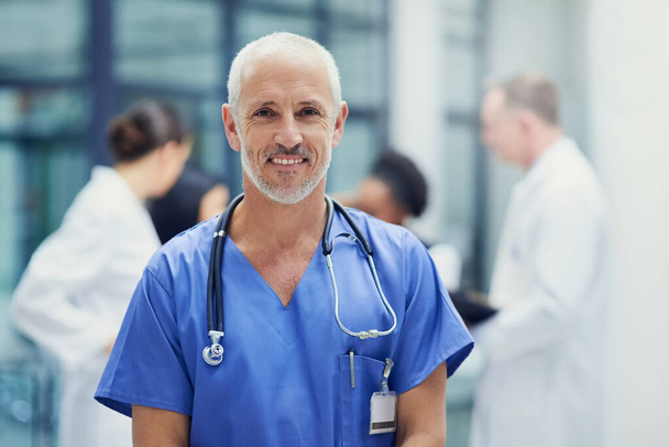 Досвід, якому можна довіряти. Портрет дорослого чоловіка-лікаря, що стоїть в лікарні з колегами на задньому плані
 - Фото, зображення