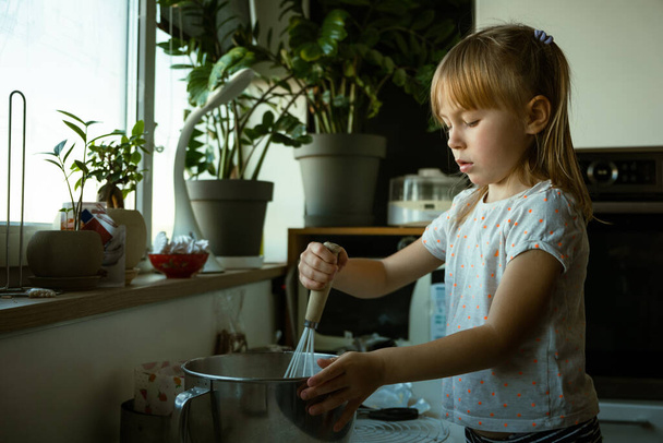 Cute little girl helping her mother to make some homemade pastry in a modern kitchen. Kid having fun while mixing ingredients in a metal bowl and cooking at home. - Zdjęcie, obraz