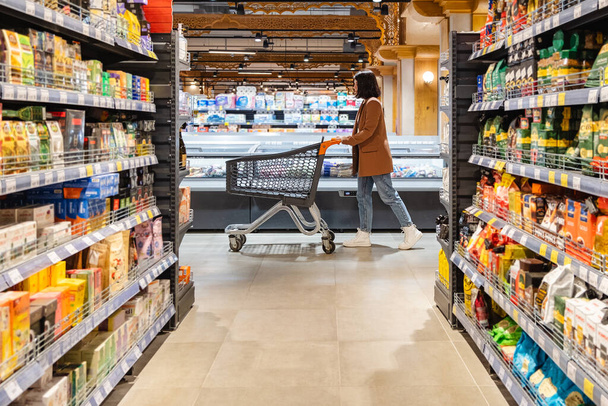 a woman with a cart walks between rows of shelves in a grocery store - 写真・画像