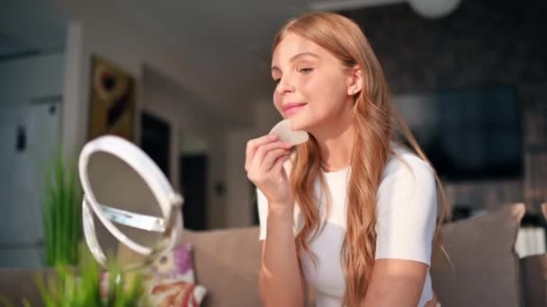 Una joven mujer caucásica está aplicando un producto de cuidado a su cara mirando a la cámara - Metraje, vídeo