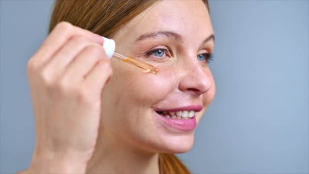 Een jonge blanke glimlachende vrouw brengt een cosmetische olie op haar gezicht aan. Langzame beweging - Video