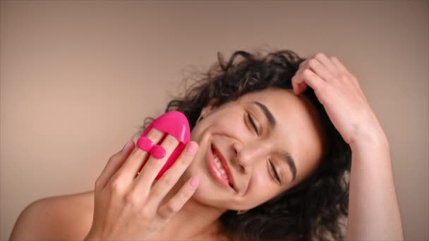 Una joven mujer caucásica sonriente está haciendo un masaje facial usando un limpiador facial, mirando a la cámara. Movimiento lento - Metraje, vídeo