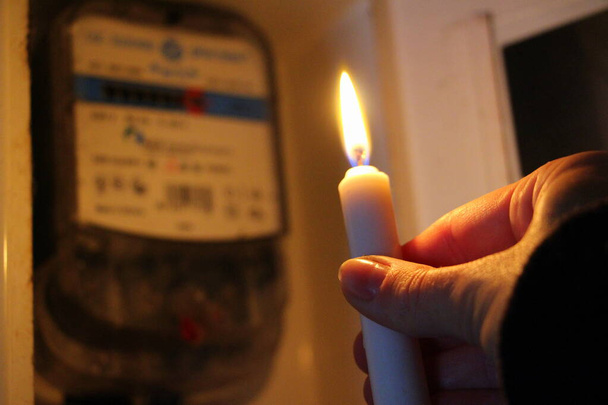 La candela in mano brilla al buio vicino al contatore dell'elettricità durante un'interruzione di corrente a casa. Città, paese senza elettricità novembre, 2022 - Foto, immagini