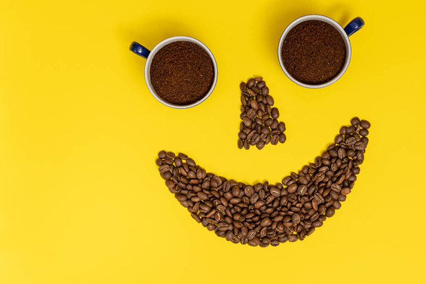 emotikon s úsměvem, rty z kávových zrn a oči z šálků kávy, složení na žlutém pozadí. - Fotografie, Obrázek