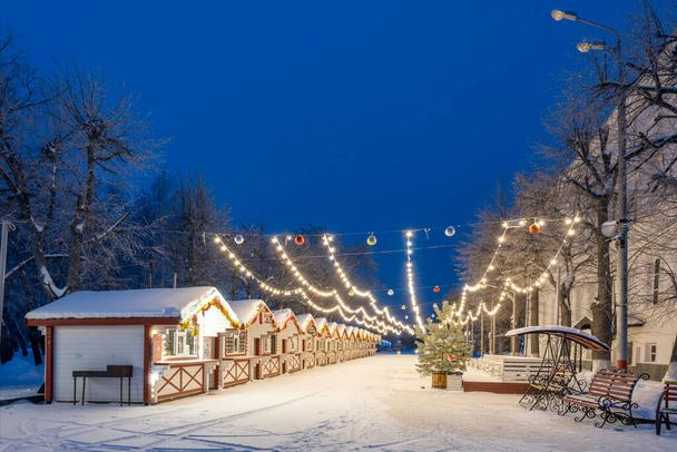 Ulyanovsk, Rússia - 14 de fevereiro de 2022: As casas de madeira de Ano Novo decoradas com grinaldas e ouropel em uma manhã gelada nevada adiantada. Rua coberta de neve em decorações de Natal. - Foto, Imagem