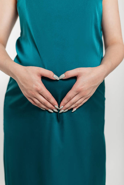 Hermosa mujer embarazada de moda en un vestido verde con un gran vientre muestra un signo de corazón con sus manos - Foto, Imagen