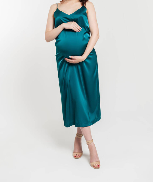 Stylish pregnant woman in a fashionable dress holds her belly and stands in the studio on a white background - Photo, Image