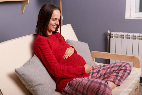 Image of young adult pregnant smiling woman wearing red jumper and pajama pants sitting on sofa in home interior, holding hands on her belly, waiting her baby. - Φωτογραφία, εικόνα
