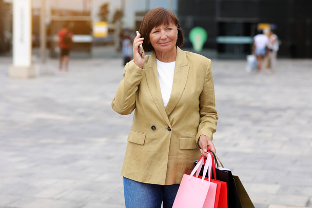 Черная пятница. Улыбающаяся женщина средних лет с кучей сумок для покупок общается по телефону через торговый центр после покупок, концепции потребления, продажи, богатой жизни. Коммерческое предложение для рекламы - Фото, изображение