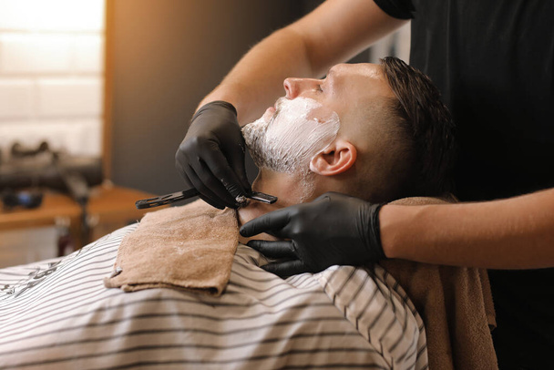Professional barber in black gloves is shaving client with a vintage straight stainless steel razor in barbershop. Shaving cream, foam. Hairdresser, beauty and fashion salon. - Foto, Imagem