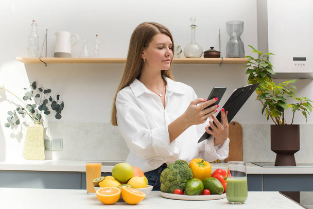 A young woman stands next to a table with fresh vegetables and fruits and makes her meal plan. Vegetarianism, healthy eating, diet. - Photo, image