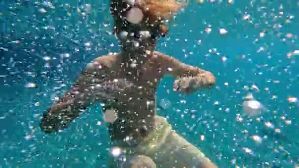 Vedenalainen teini-ikäinen poika suojalaseissa sukeltaa kirkkaaseen siniseen veteen, jota ympäröivät valkoiset ilmakuplat ja heiluttavat kättään. - Materiaali, video