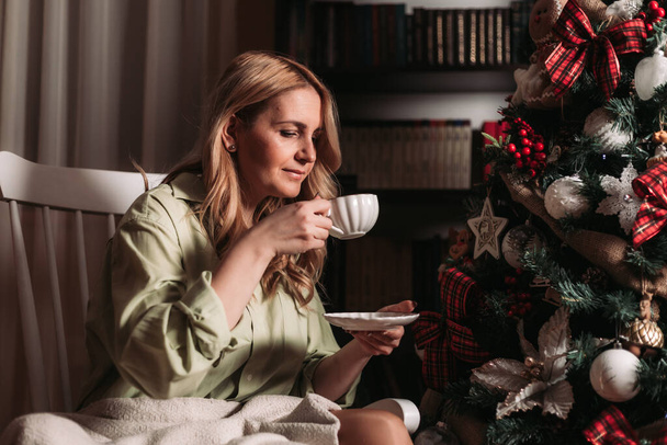 Young blonde woman sitting on a rocking chair next to a Christmas tree drinking tea - Fotó, kép