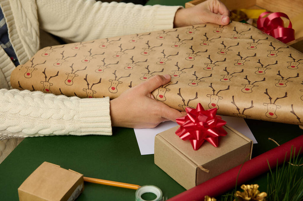 装飾的な素材を背景にクリスマスの贈り物を詰めながら、鹿のパターンで包装紙を展開する女性の手のクローズアップ。ボクシングの日だ。死の贈り物だ。冬のイベント - 写真・画像