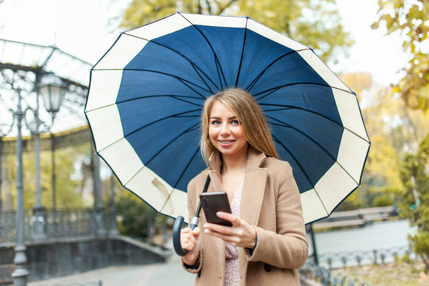 Fashion vrouw in een stijlvolle jas met paraplu via telefoon en wandelingen door het herfstpark - Foto, afbeelding