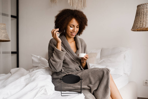 African american woman in bathrobe with facial moisturizing cream doing morning beauty routine in bedroom. African girl applying face moisturizer cream to protect skin from dryness - 写真・画像