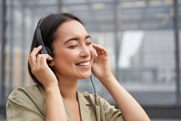 Egy gyönyörű ázsiai nő portréja fülhallgatóban, zenét hallgatva a belváros utcáin, boldogan mosolyogva. - Fotó, kép
