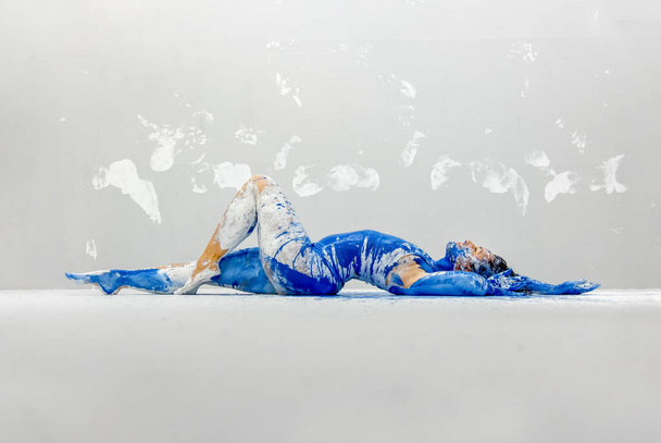 mladá sexy žena ve spodním prádle, sportovní oblečení, umělecky abstraktní malováno bílou a modrou barvou, ležící na podlaze v ateliéru umělců, stěna s bílými potisky, kopírovací prostor. - Fotografie, Obrázek