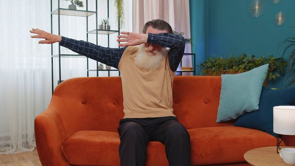 Щасливий задоволений старший бородатий чоловік танцює на дивані вдома, вибираючи слухати улюблену енергійну диско-музику, роблячи жест даб. Літній дідусь танцюрист. Люди вихідні розважальні заходи
 - Фото, зображення