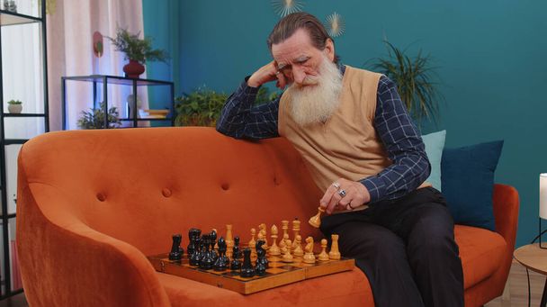 Сосредоточенный старший дедушка шахматист, сидящий на домашнем диване и обучающийся игре в шахматы в одиночку, воспитательной досуговой домашней деятельности, стратегическим навыкам. Пожилой пенсионер - Фото, изображение