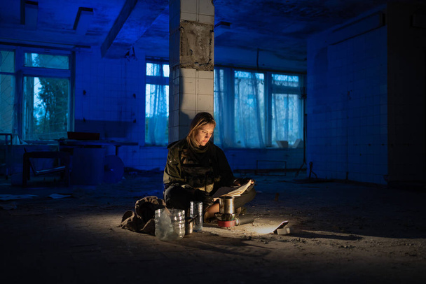 sale femme fatiguée assis sur le sol dans une chambre abandonnée la nuit lit un journal brille avec une lanterne. Après l'apocalypse. randonnées dans un monde post-apocalyptique. espace de copie. - Photo, image