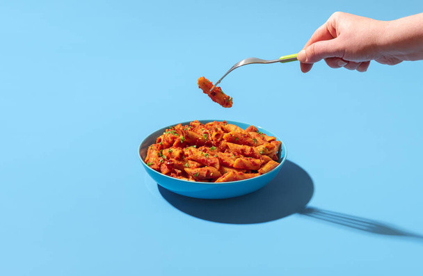 Woman's hand taking a pasta tube from a bowl with a fork. Eating pasta penne alla arrabbiata. Spicy pasta dish in a blue bowl minimalist on a colorful table. - Photo, Image