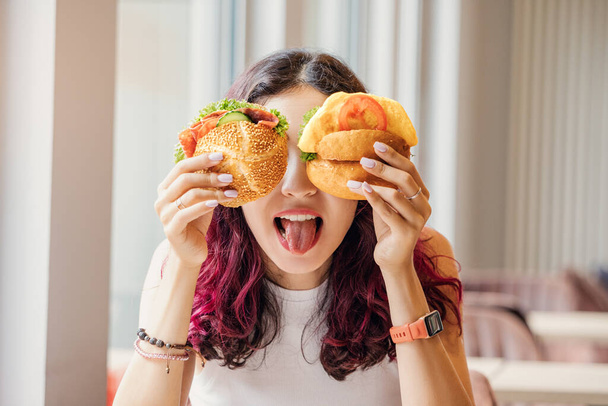 A girl in a cafe fooling around and closed her eyes with two cheeseburgers. Fast food and healthy diet concept - Photo, Image