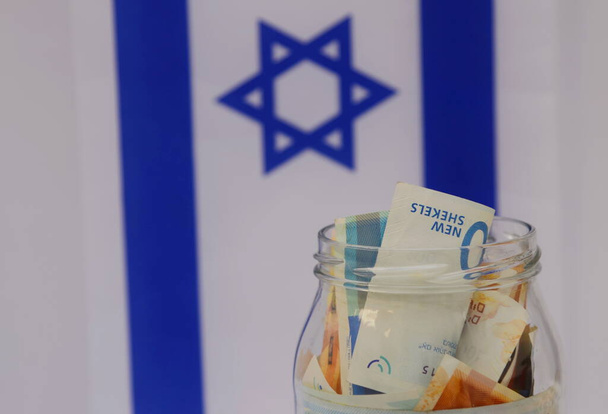 Glazen pot met Israëlische sikkel op de achtergrond van de vlag van Israël. Begrip: persoonlijk sparen, bankwezen, inflatie, nationale munt - Foto, afbeelding