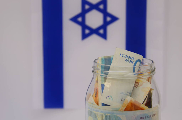 Üvegedény izraeli sékellel Izrael zászlajának hátterében. Koncepció: személyes megtakarítások, bankrendszer, infláció, nemzeti valuta - Fotó, kép