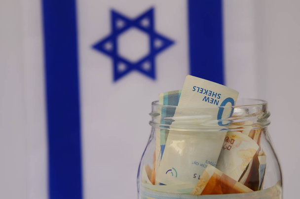 Pot en verre avec shekel israélien sur le fond du drapeau d'Israël. Concept : épargne personnelle, système bancaire, inflation, monnaie nationale - Photo, image