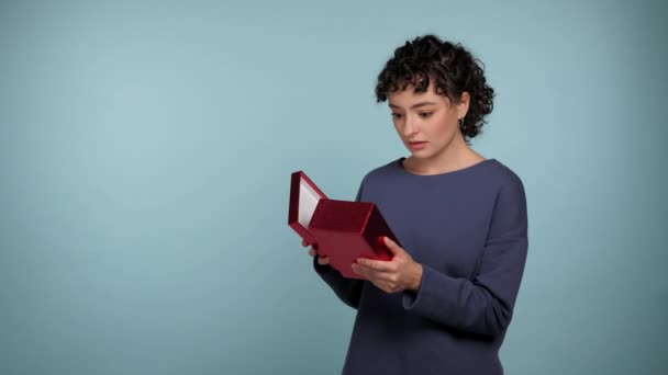 Zszokowany kręcone kobieta otwiera prezent czerwone pudełko zaskoczony wygląda aparat fotograficzny i mówi wow. Podekscytowana kobieta nosząca niebieski sweter z prezentem w dłoniach na odizolowanym jasnoniebieskim tle z przestrzenią do kopiowania - Materiał filmowy, wideo