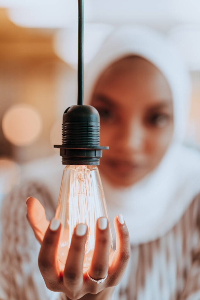 Sentimental portrait of African-American young female Muslim wearing hijab while holding a lightbulb - 写真・画像