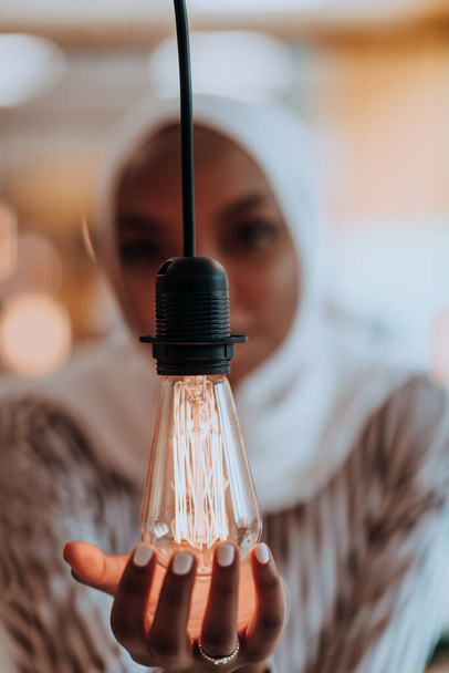 Sentimental portrait of African-American young female Muslim wearing hijab while holding a lightbulb - Φωτογραφία, εικόνα