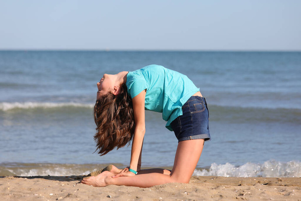 junges schlankes Mädchen in blauem T-Shirt und kurzer Jeanshose turnt im Sommer am Meer - Foto, Bild
