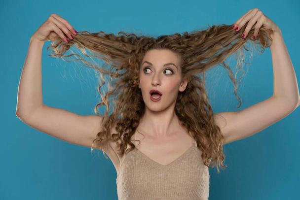 Attractive amazed blonde woman showing her curly beautiful hair  on blue studio background - Photo, image