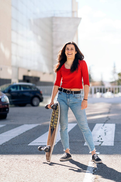 Happy young woman with casual clothes standing in zebra crossing holding her long-board and looking at camera. - Foto, imagen