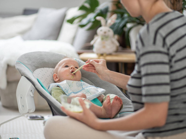 Mère cuillère nourrir son bébé garçon enfant dans une chaise bébé avec de la purée de fruits. Concept d'introduction aux aliments solides pour bébés - Photo, image