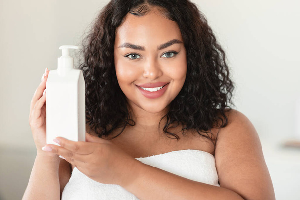 Piękny czarny plus size kobieta trzyma butelkę z balsamem nawilżającym do ciała, przy użyciu nowego produktu do pielęgnacji skóry po kąpieli, uśmiechnięty do kamery, przestrzeń do kopiowania - Zdjęcie, obraz
