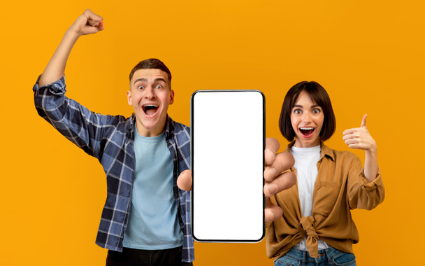 Happy excited millennial european couple celebrate victory with hands gesture, open mouth, show big phone with blank screen, isolated on orange background. Online victory, new app and people emotions - Foto, Imagen