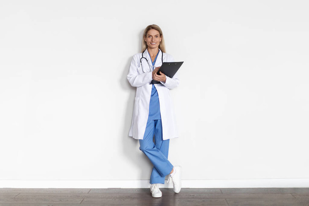 Портрет улыбающейся женщины-врача в униформе с буфером в руках, стоящих рядом с белой стеной в помещении, счастливый врач леди в медицинском пальто и стетоскоп глядя на камеру, скопировать пространство - Фото, изображение
