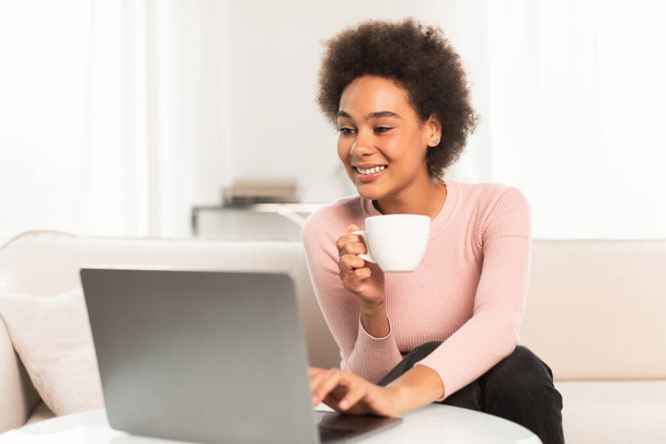 Heureuse femme afro-américaine millénaire profiter d'une tasse de café et dactylographier sur ordinateur portable dans un salon intérieur lumineux. Travail à distance, freelance, appel vidéo, mode de vie et distance sociale à la maison, covid-19 - Photo, image