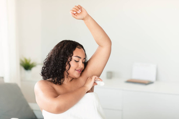 Mladá černošky tělo pozitivní žena pomocí antiperspirant tyčinka na podpaží, dáma použití deodorant na podpaží pro prevenci pocení, sedí v ložnici, volný prostor - Fotografie, Obrázek