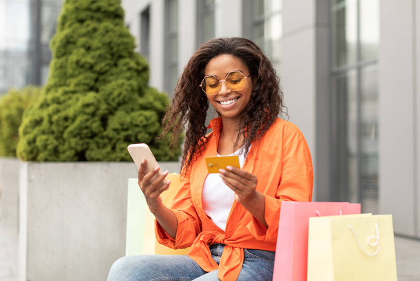 Cheerful millennial black female in glasses with many bags reads message on smartphone, buy purchases, holds credit card in city. Offer, ad and huge sale for online shopping, lifestyle and finance - Valokuva, kuva