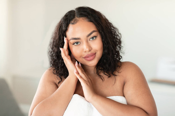Pretty black overweight lady enjoying perfect soft skin after spa procedures, posing wrapped in towel at home. Body care, treatment and natural beauty - 写真・画像