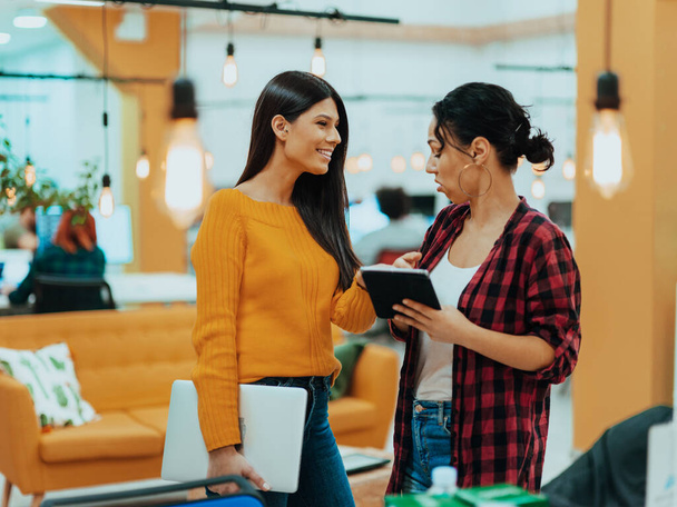 Portrait of two businesswomen talking to each other while standing in a modern business office with their colleagues using laptops and tablets. Marketing concept. Multi-ethnic society - Photo, Image