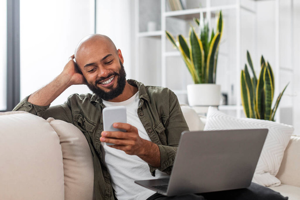 Positive mature latin male freelancer sitting on sofa with laptop and using cellphone, texting online while working from home, looking at phone screen and smiling - 写真・画像