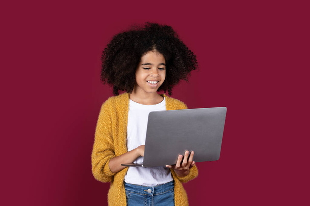 Adorable black girl with bushy hair schooler using modern computer, typing on laptop keyboard and smiling, preteen kid doing homework online, isolated on burgundy studio background, copy space - Photo, Image