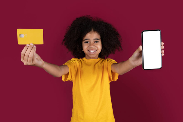 Adorable preteen black girl with bushy hair in yellow t-shirt showing modern smartphone with white blank screen and yellow plastic credit card, burgundy studio background, mockup. Online shopping - Photo, Image