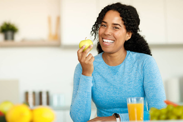 Portrait of excited black woman in sportswear holding apple and smiling at camera, sitting in light kitchen interior. Healthy eating and nutrition for slimming, low calories food recipes - Photo, image
