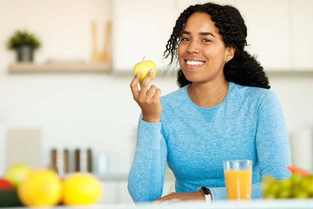 Nutrición para perder peso. Feliz chica de fitness negro sentado con manzana y sonriendo a la cámara, sentado en la cocina, espacio libre. Dieta saludable para adelgazar. Cocinar para el concepto de pérdida de peso - Foto, imagen
